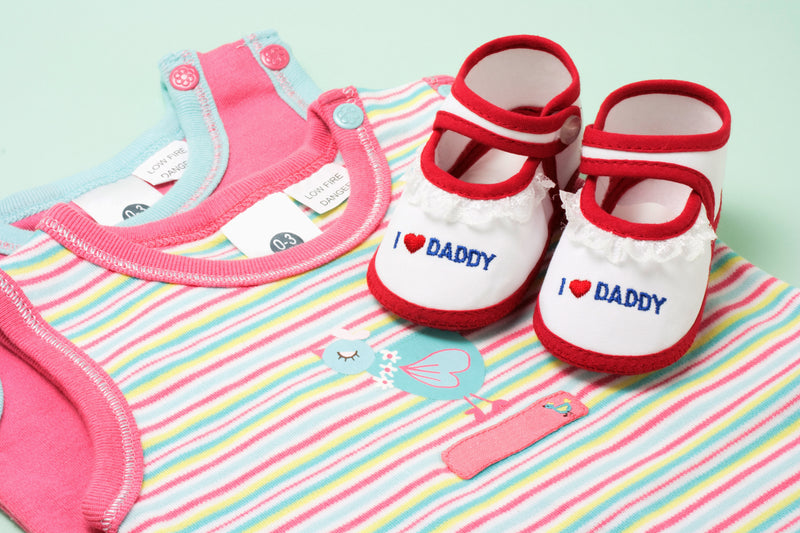 Baby clothing set with pink bib, striped shirt, and red and white shoes on a colorful blanket.