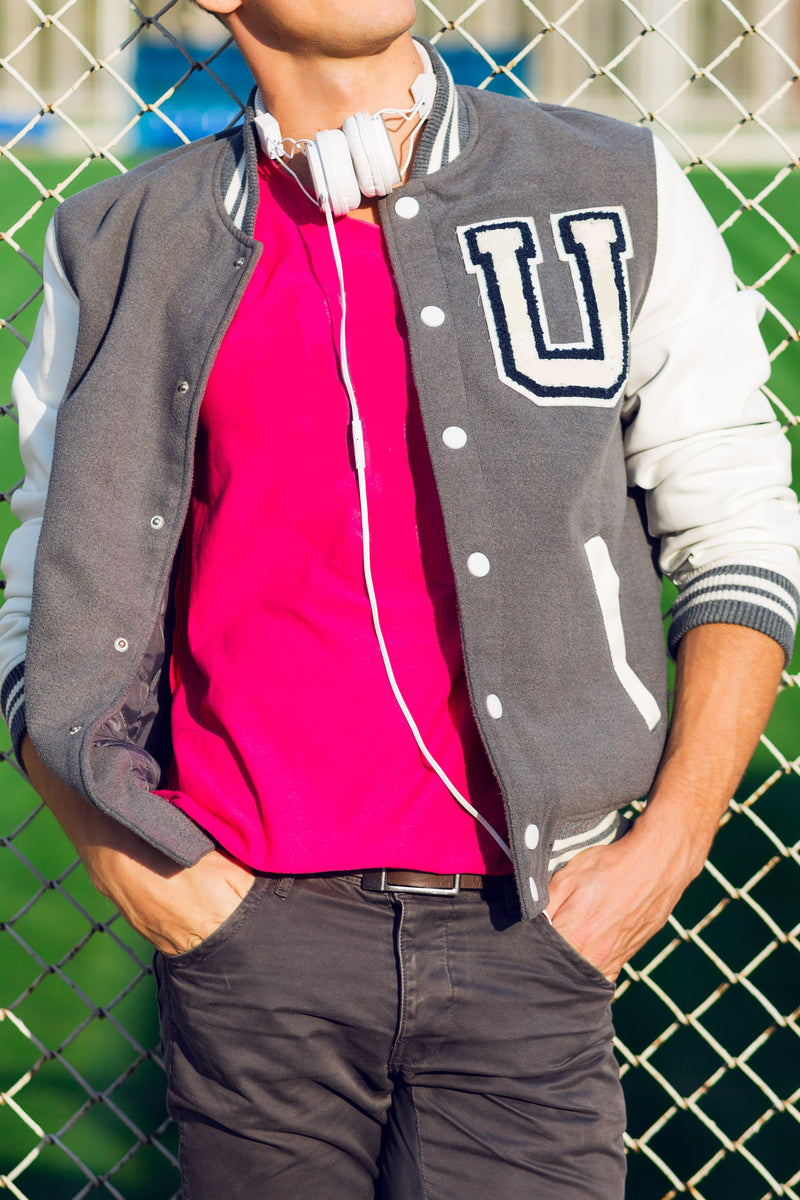 Person wearing a gray varsity jacket with a letter 'U' on a chain-link fence background